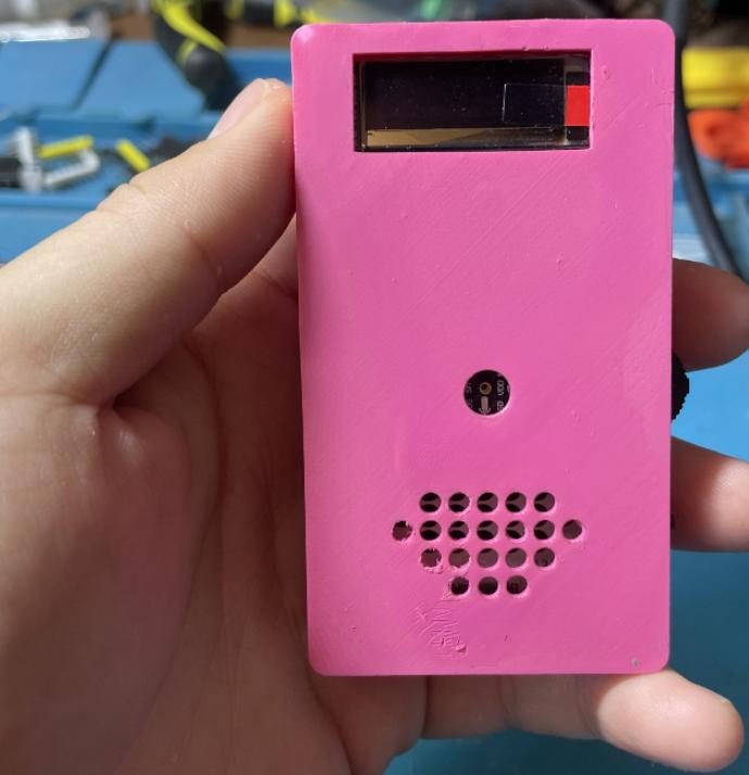Hand holding a pink 3D-printed rectangular device with a small screen, a microphone hole and a speaker grille, over a cluttered workbench