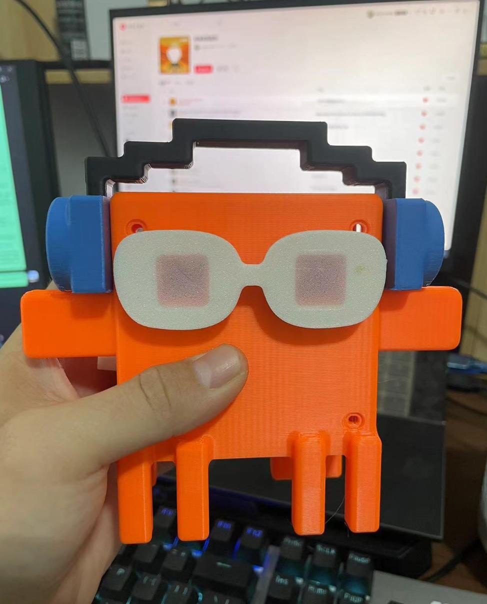 Hand holding an orange 3D-printed character with white glasses and blue headphones in front of a computer monitor and keyboard