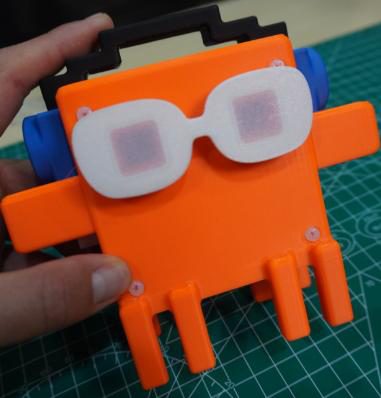 Hand holding the orange 3D-printed character above a green cutting mat