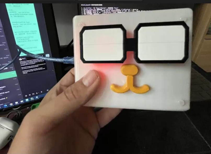 Hand holding a white square box with black glasses and a yellow moustache, a red light glowing, in front of a monitor