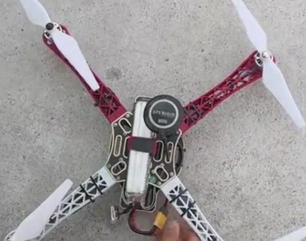 Top view of an assembled quadcopter with red and white arms, white propellers, a GPS puck and a strapped battery, on concrete ground