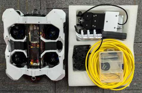 White flat-frame ROV seen from above on a grey carpet, next to a foam case holding the gripper, yellow tether cable and a small clear parts box