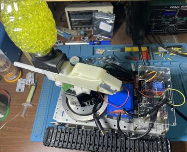 High-angle view of the tracked robot on a blue workbench mat, with the yellow gel-bead hopper on the launcher and tools around
