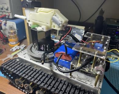Angled view of the tracked robot's turret, battery and acrylic electronics stack in front of bench equipment
