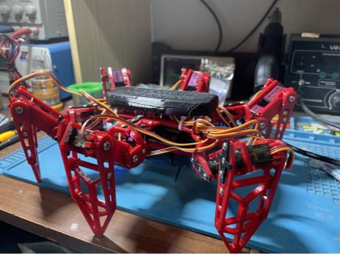 Side view of the red hexapod standing on a blue anti-static mat on a workbench, with a power supply and soldering station behind it
