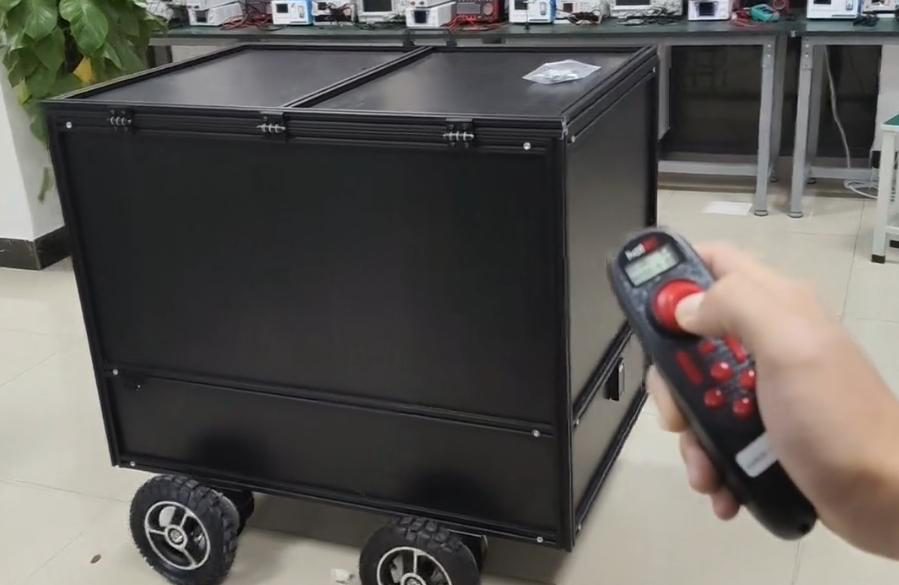 Black aluminium-frame cargo robot on a lab floor, a hand in the foreground pointing a handheld remote at it, benches with test instruments and a potted plant behind