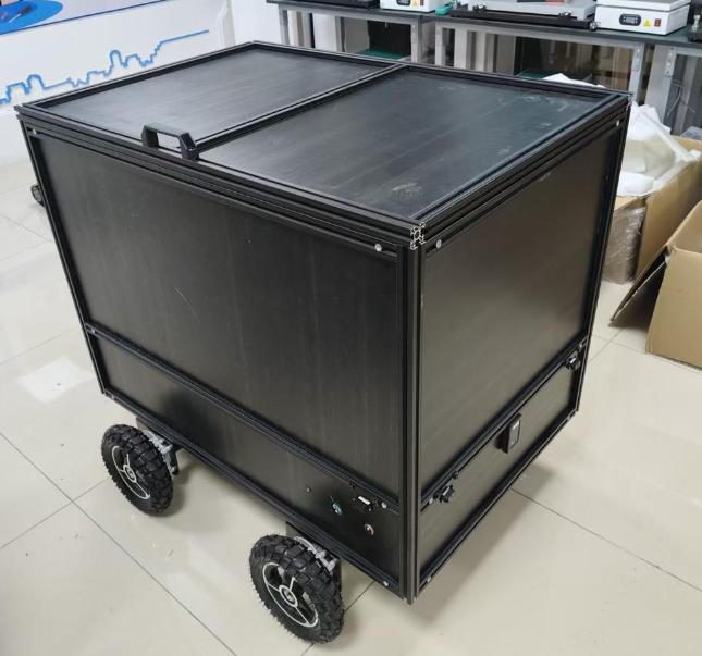 Black box-shaped delivery robot with a split lid and four air-filled wheels on a tiled floor, cardboard boxes and lab benches behind