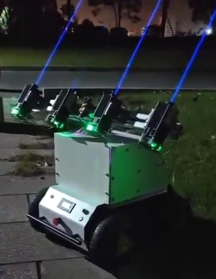 White four-wheel robot outdoors at night on paving beside grass, four laser modules on a beam above it firing blue beams into the sky, green status lights