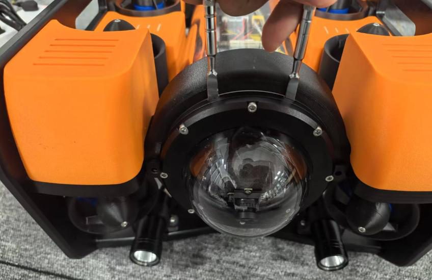 Front of an ROV with orange thruster shells on a grey carpet: clear camera dome in a black housing, two small lights below, a hand holding two metal rods above