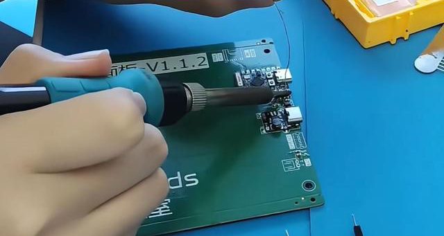 Hand soldering a component onto a green PCB on a blue anti-static mat