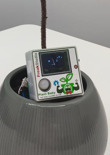 Close-up of the square device in the pot showing a small screen, a round motion sensor and a cartoon plant label