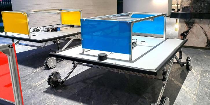 Two large flat-deck robots with blue and yellow acrylic side panels on aluminium frames, mecanum wheels and spring suspension, parked on a dark marble floor in a lobby, a lidar puck on the deck
