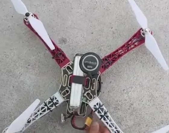 Top view of an assembled quadcopter with red and white arms, white propellers, a GPS puck and a strapped battery, on concrete ground