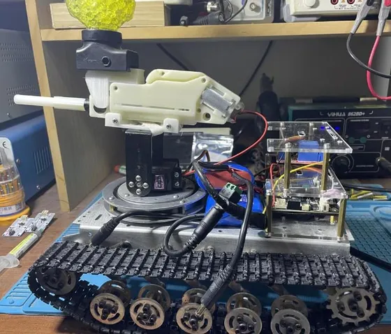 Tracked metal chassis with a white 3D-printed launcher on a servo turret, blue battery pack and acrylic electronics enclosure, on a workbench with a hopper of yellow gel beads and a bench power supply