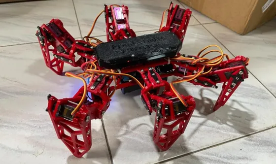 Red 3D-printed six-legged robot with a black body cover standing on a tiled floor next to a cardboard box