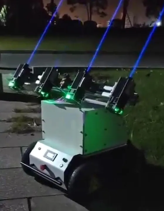 White four-wheel robot outdoors at night on paving beside grass, four laser modules on a beam above it firing blue beams into the sky, green status lights