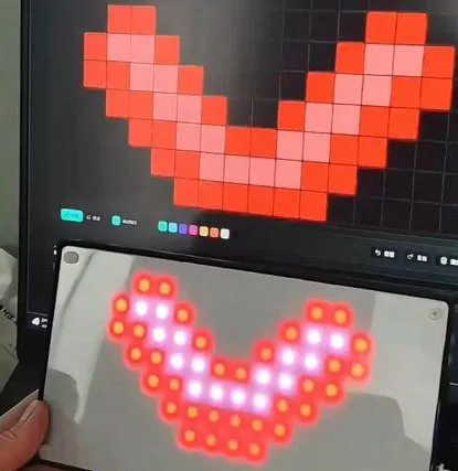 Hands holding a lit LED panel showing a red pixel heart shape in front of a screen displaying the same shape in an editor