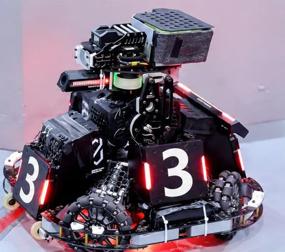Black competition robot with number '3' armor plates lit by red LED strips, omni wheels and a gimbal on top, against a pink wall