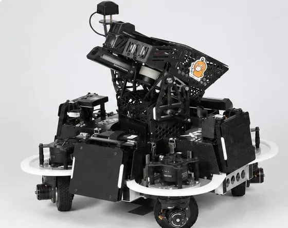 Black carbon-fiber competition robot with white bumper rings and a tilted gimbal on a grey studio backdrop; each wheel sits under its own steering motor