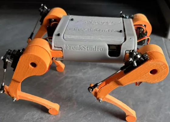 Small orange-and-grey 3D-printed quadruped robot standing on a dark grey surface