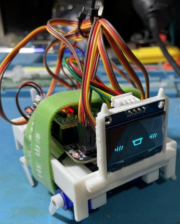 Small white 3D-printed quadruped robot with an OLED face, a green strap and many servo wires, on a blue workbench mat