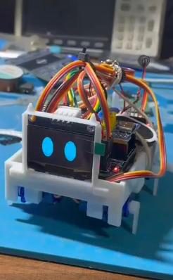 Front view of the small robot with two blue eyes on its OLED screen, on a blue mat with test equipment behind