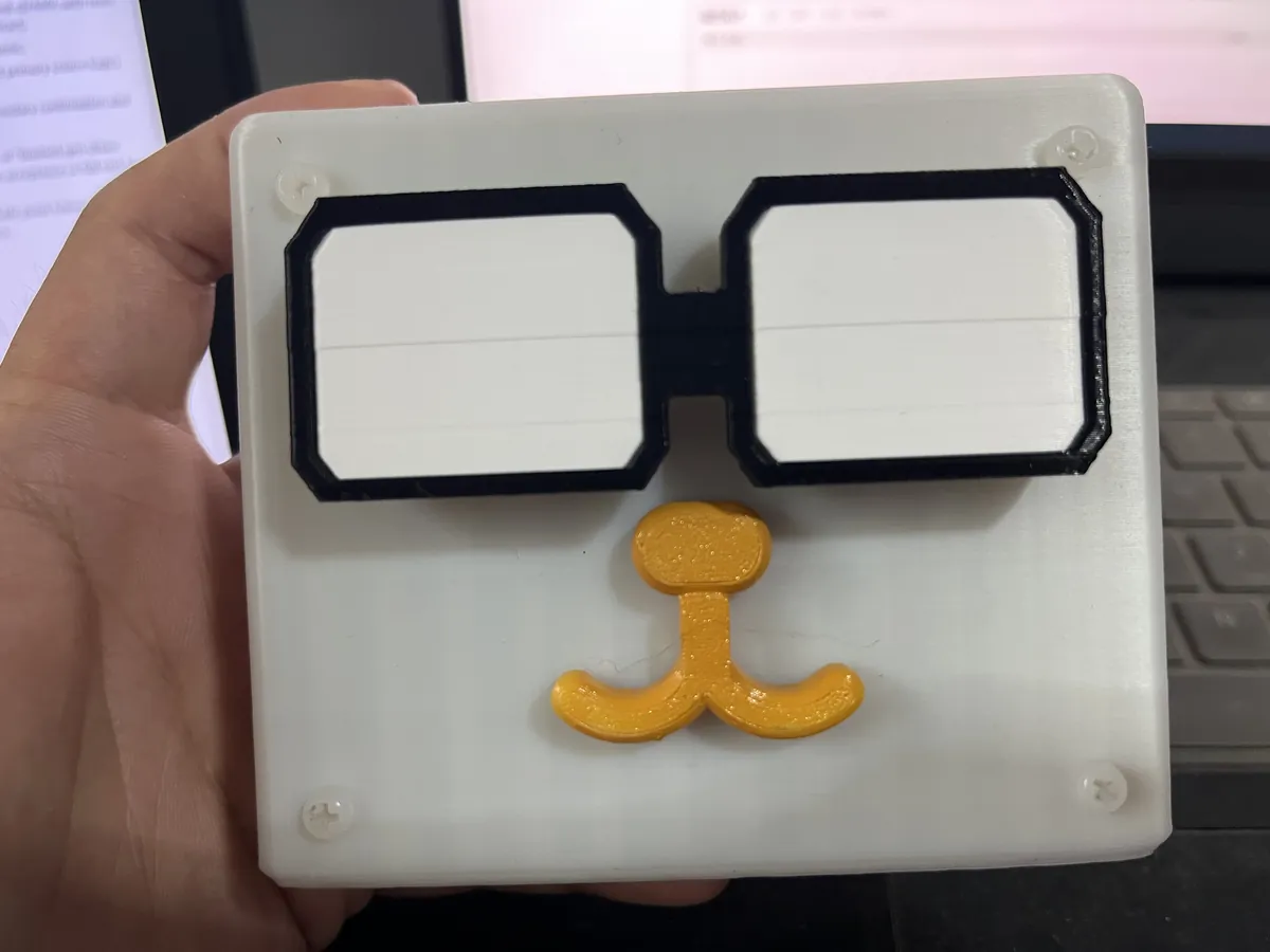 Hand holding a white 3D-printed square enclosure with two glasses-shaped light windows and an orange mustache on the front