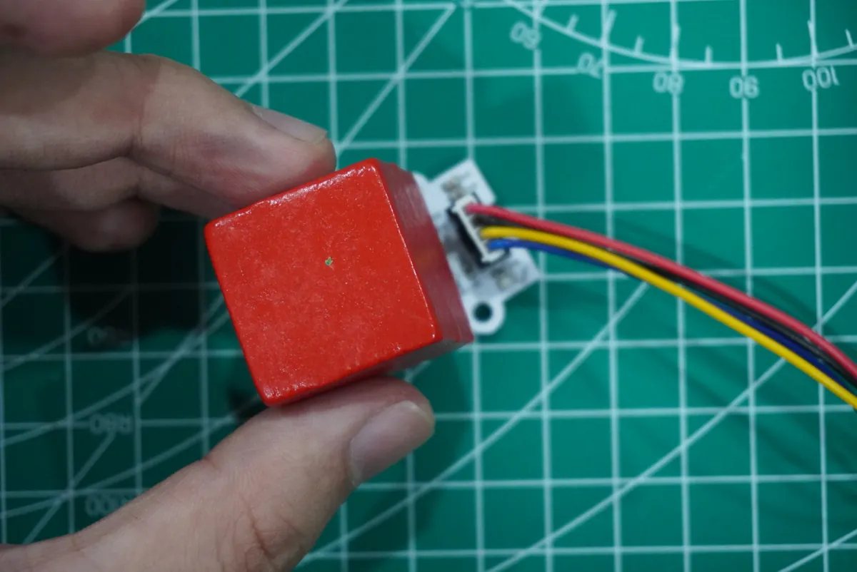 Hand holding a red wooden cube against a small color sensor board with a four-wire cable on a cutting mat