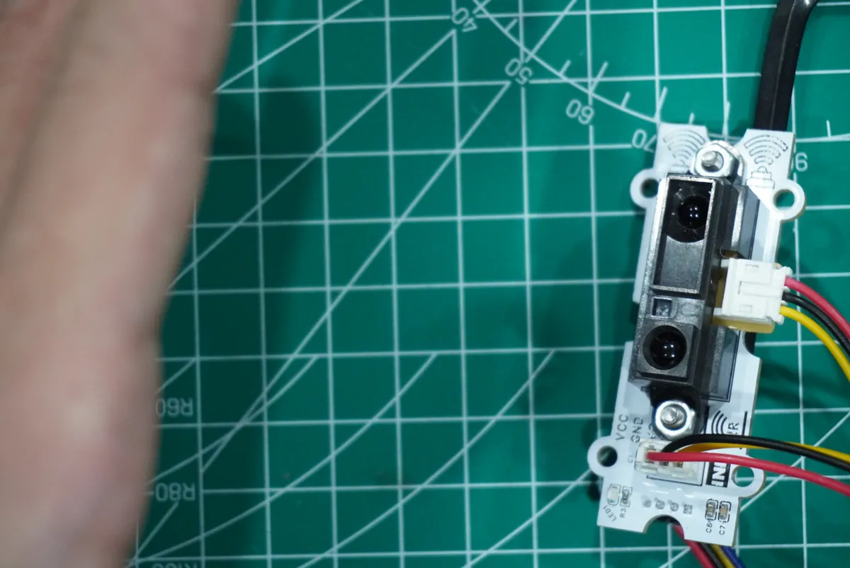 A hand held to the left of a Sharp analog infrared distance sensor module on a green cutting mat