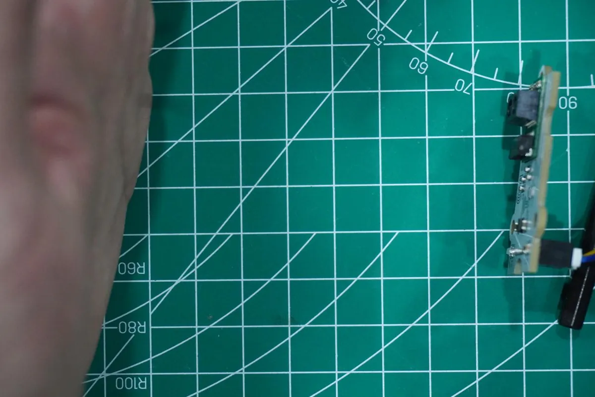 A hand held to the left of a small digital infrared distance module mounted on edge on a green cutting mat