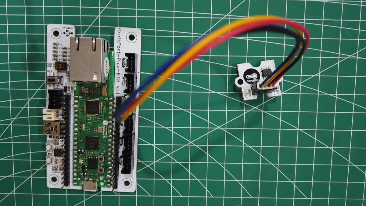 A small temperature sensor module connected by a four-wire cable to a GraftPort-Pico-ETH board on a cutting mat