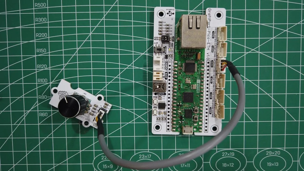 Rotary encoder module with a black knob connected by a gray cable to a GraftPort-Pico-ETH carrier board on a cutting mat