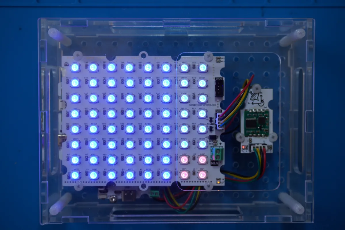 Clear acrylic case holding an 8 by 8 LED matrix lit blue with a 2 by 2 red block in one corner, and a motion sensor module beside it